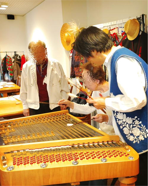 青いハンガリー刺繍のベストを着た奏者 斉藤浩が、参加者の手元にバチを添えてツィンバロンの弾き方を指導している。テーブルには麦わら帽子と衣装が並ぶ。